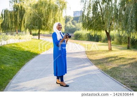 An African American Muslim woman wearing a hijab and a blue dress holds a camera while walking on a path in a park An African American Muslim woman wearing a hijab and a blue dress holds a camera while walking on a path in a park 142630644