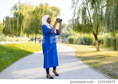 An African American Muslim woman in a blue dress and hijab holds a camera to take pictures in a park An African American Muslim woman in a blue dress and hijab holds a camera to take pictures in a park 142630674