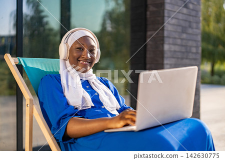 A happy African American Muslim woman wearing a hijab and headphones works on her laptop outdoors while relaxing on a lounge chair 142630775