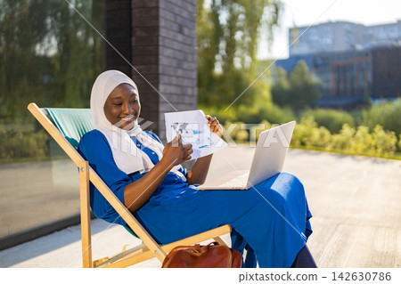 A smiling African American Muslim woman wearing a hijab and blue dress works on her laptop and reviews documents while relaxing on a lounge chair outside A smiling African American Muslim woman wearing a hijab and blue dress works on her laptop and reviews documents while relaxing on a lounge chair outside 142630786