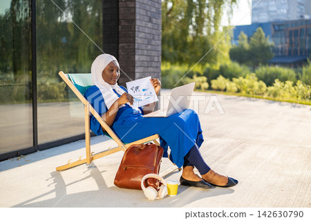 An African American Muslim woman in a hijab works on her laptop while sitting on a lounge chair outside a modern building 142630790