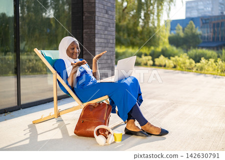 A smiling African woman wearing a hijab is engaged in a video call on her laptop while relaxing on a lounge chair outside 142630791