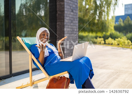 A happy African American Muslim woman wearing a hijab is enjoying a video call on her laptop while relaxing on a lounge chair outside A happy African American Muslim woman wearing a hijab is enjoying a video call on her laptop while relaxing on a lounge chair outside 142630792
