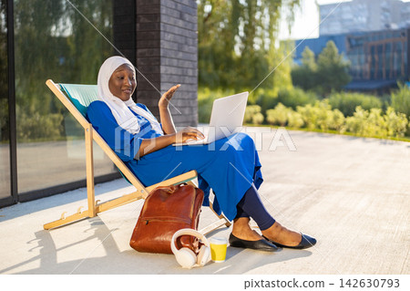 A Muslim woman wearing a hijab is working on her laptop while sitting on a lounge chair outdoors with a backpack and headphones nearby 142630793