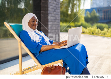 A smiling African American Muslim woman wearing a hijab and blue dress works on her laptop while relaxing on a lounge chair outside 142630795