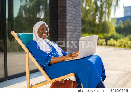 A smiling African American Muslim woman wearing a hijab and blue dress relaxes on a lounge chair with her laptop in a sunny outdoor setting A smiling African American Muslim woman wearing a hijab and blue dress relaxes on a lounge chair with her laptop in a sunny outdoor setting 142630796