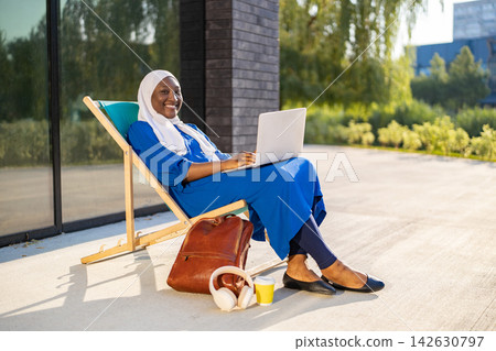 A smiling African American Muslim woman wearing a hijab and blue dress sits on a lounge chair with a laptop and backpack A smiling African American Muslim woman wearing a hijab and blue dress sits on a lounge chair with a laptop and backpack 142630797