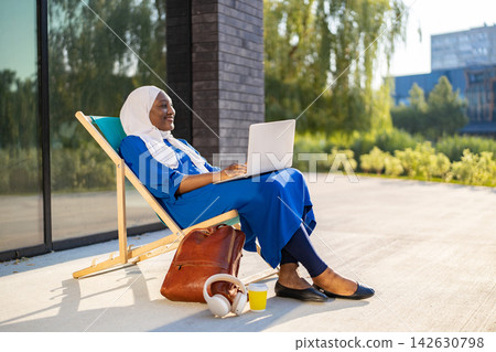 A smiling African American Muslim woman in a hijab and blue dress works on her laptop while relaxing on a lounge chair outdoors A smiling African American Muslim woman in a hijab and blue dress works on her laptop while relaxing on a lounge chair outdoors 142630798