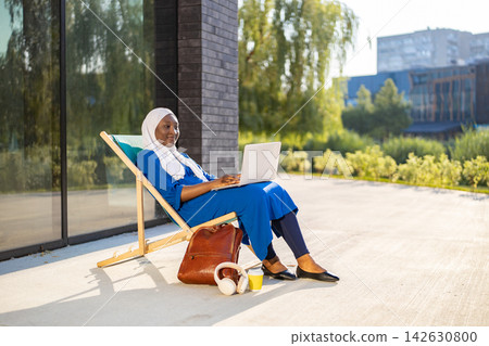 A smiling African American Muslim woman wearing a hijab works on her laptop while relaxing on a lounge chair outdoors A smiling African American Muslim woman wearing a hijab works on her laptop while relaxing on a lounge chair outdoors 142630800