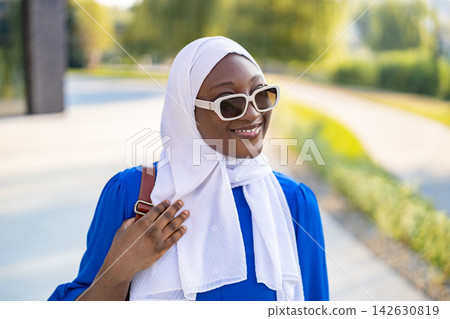 A young African American Muslim woman wearing a white hijab and sunglasses smiles while walking outdoors on a sunny day A young African American Muslim woman wearing a white hijab and sunglasses smiles while walking outdoors on a sunny day 142630819