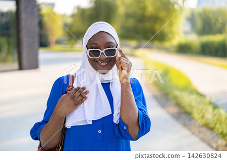 A happy African American Muslim woman wearing a white hijab and sunglasses poses outdoors with a bright smile A happy African American Muslim woman wearing a white hijab and sunglasses poses outdoors with a bright smile 142630824