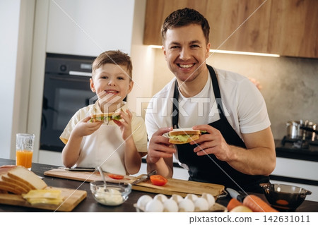Caucasian father and son smiling while holding sandwiches in a modern kitchen with ingredients and cooking utensils on the countertop Caucasian father and son smiling while holding sandwiches in a modern kitchen with ingredients and cooking utensils on the countertop 142631011