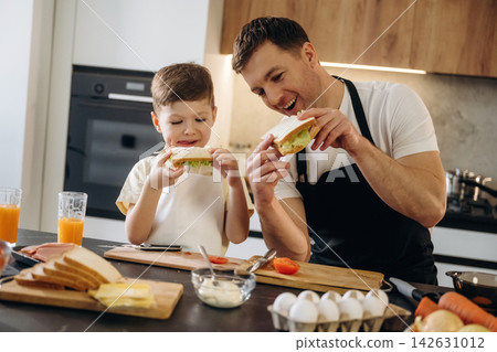 Father and son preparing sandwiches together in a modern kitchen with fresh ingredients, including bread, vegetables, and drinks on the countertop 142631012