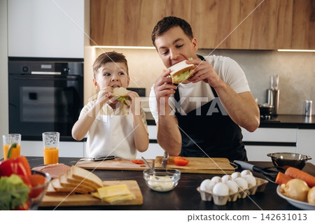 Father and son enjoying homemade sandwiches together in a modern kitchen with fresh ingredients and cooking utensils on the countertop Father and son enjoying homemade sandwiches together in a modern kitchen with fresh ingredients and cooking utensils on the countertop 142631013