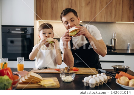Father and son enjoying sandwiches together in a modern kitchen, surrounded by fresh vegetables, eggs, and kitchen utensils on wooden countertops Father and son enjoying sandwiches together in a modern kitchen, surrounded by fresh vegetables, eggs, and kitchen utensils on wooden countertops 142631014