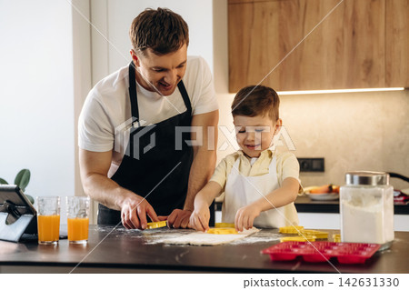Father and son baking cookies together in a modern kitchen, using cookie cutters on a floured countertop with orange juice glasses nearby Father and son baking cookies together in a modern kitchen, using cookie cutters on a floured countertop with orange juice glasses nearby 142631330