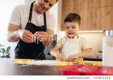Adult male and young boy in aprons preparing Easter cookies together in a modern kitchen, with cookie cutters and flour spread across the countertop Adult male and young boy in aprons preparing Easter cookies together in a modern kitchen, with cookie cutters and flour spread across the countertop 142631339