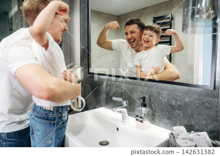 Father and son flexing muscles in front of bathroom mirror, smiling joyfully while standing by sink with soap dispenser and towels visible in modern interior design Father and son flexing muscles in front of bathroom mirror, smiling joyfully while standing by sink with soap dispenser and towels visible in modern interior design 142631382