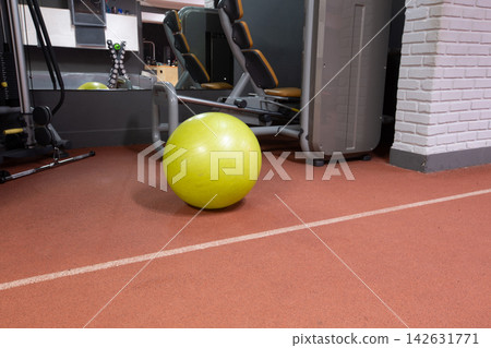 Yellow exercise ball sits on gym floor near workout equipment during indoor training session Yellow exercise ball sits on gym floor near workout equipment during indoor training session 142631771