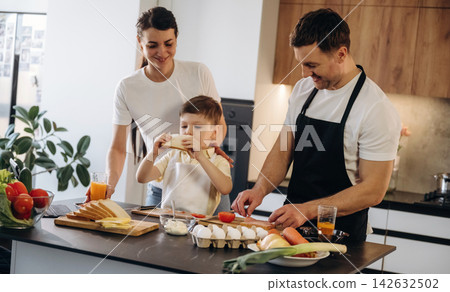Family preparing breakfast together in a modern kitchen with fresh ingredients, including bread, eggs, and vegetables on the countertop Family preparing breakfast together in a modern kitchen with fresh ingredients, including bread, eggs, and vegetables on the countertop 142632502