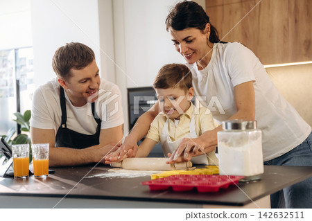 Family cooking together in a modern kitchen, mother and father assisting young boy with rolling dough on countertop surrounded by ingredients and kitchenware Family cooking together in a modern kitchen, mother and father assisting young boy with rolling dough on countertop surrounded by ingredients and kitchenware 142632511