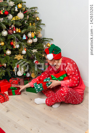 A 52-year-old woman sits on the floor next to a Christmas tree. She wears festive clothes and holds a decoration as she looks at gifts on December 25th. The room is filled with holiday cheer A 52-year-old woman sits on the floor next to a Christmas tree. She wears festive clothes and holds a decoration as she looks at gifts on December 25th. The room is filled with holiday cheer 142633201