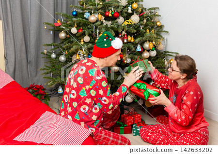 A couple is smiling and exchanging gifts on Christmas Day. They are in a cozy room, sitting by a Christmas tree full of decorations. The festive atmosphere is filled with joy and celebration A couple is smiling and exchanging gifts on Christmas Day. They are in a cozy room, sitting by a Christmas tree full of decorations. The festive atmosphere is filled with joy and celebration 142633202