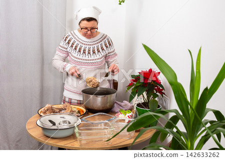 A woman stands in a kitchen, focusing on preparing aspic, kholodets, a traditional dish. She removes boiled meat from a pot with broth while gathering utensils and containers for the meal A woman stands in a kitchen, focusing on preparing aspic, kholodets, a traditional dish. She removes boiled meat from a pot with broth while gathering utensils and containers for the meal 142633262