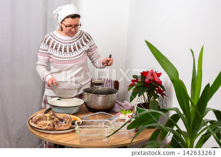 A woman stands in her kitchen preparing kholodets, aspic, a traditional Ukrainian dish. She strains the broth from the bones into a bowl while wearing comfortable clothes A woman stands in her kitchen preparing kholodets, aspic, a traditional Ukrainian dish. She strains the broth from the bones into a bowl while wearing comfortable clothes 142633263