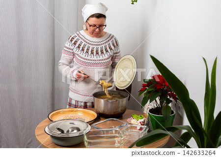 A woman stands in a kitchen, focusing on preparing aspic, kholodets, a traditional dish. She removes boiled meat from a pot with broth while gathering utensils and containers for the meal 142633264
