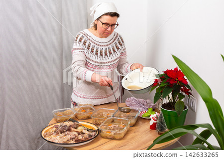 A woman stands by a table in a kitchen, preparing kholodets, aspic, or jellied meat, traditional Ukraine dish. She carefully pours broth from a large bowl into glass containers A woman stands by a table in a kitchen, preparing kholodets, aspic, or jellied meat, traditional Ukraine dish. She carefully pours broth from a large bowl into glass containers 142633265