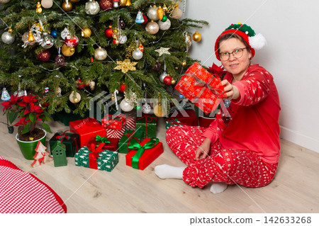 On Christmas Day, a 52-year-old woman sits on the floor near a Christmas tree. She smiles while holding wrapped gifts and surrounded by decorations in her cozy living room On Christmas Day, a 52-year-old woman sits on the floor near a Christmas tree. She smiles while holding wrapped gifts and surrounded by decorations in her cozy living room 142633268