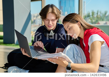 Two young students studying with laptop and notebook sitting on floor near big window 142633496