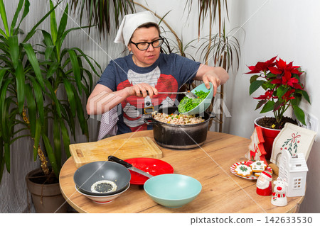 Woman prepares Olivier salad, Russian salad, a traditional Ukrainian or Russian Christmas or New Year dish in a kitchen. She adds boiled green peas to a bowl with other ingredients 142633530