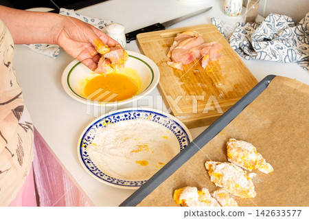 In a kitchen, a woman dips chicken breast pieces in egg while preparing to make deep-fried chicken nuggets. Flour sits on a plate beside her In a kitchen, a woman dips chicken breast pieces in egg while preparing to make deep-fried chicken nuggets. Flour sits on a plate beside her 142633577
