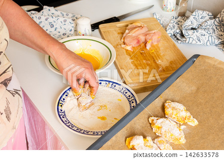 A woman is deep-frying chicken nuggets at her kitchen. She dips breast pieces in flour. The environment shows a clean counter with raw chicken and a bowl of egg A woman is deep-frying chicken nuggets at her kitchen. She dips breast pieces in flour. The environment shows a clean counter with raw chicken and a bowl of egg 142633578