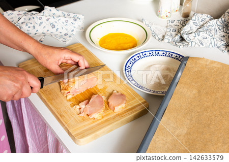 A woman cuts chicken on a wooden board with a knife. Nearby, there is a bowl of beaten eggs and a plate with flour, all set for making deep-fried chicken nuggets A woman cuts chicken on a wooden board with a knife. Nearby, there is a bowl of beaten eggs and a plate with flour, all set for making deep-fried chicken nuggets 142633579