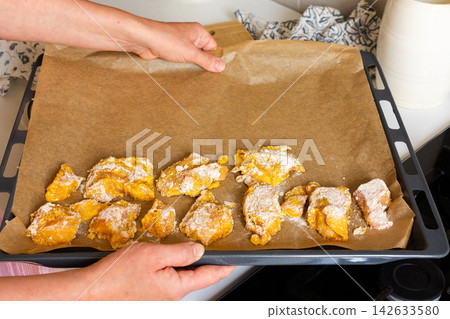 Hands hold a baking tray with deep-fried chicken nuggets covered in flour. The scene is set in a home kitchen with light colors and a casual atmosphere 142633580