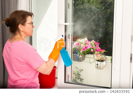 A woman in orange gloves sprays cleaner on a window as she stands beside a bucket. Bright flowers in pots are visible outside on the balcony during daylight A woman in orange gloves sprays cleaner on a window as she stands beside a bucket. Bright flowers in pots are visible outside on the balcony during daylight 142633591
