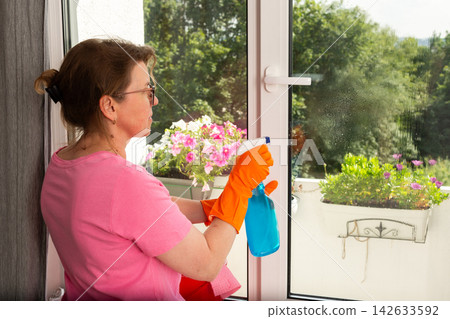 A woman in orange gloves sprays cleaner on a window as she stands beside a bucket. Bright flowers in pots are visible outside on the balcony during daylight A woman in orange gloves sprays cleaner on a window as she stands beside a bucket. Bright flowers in pots are visible outside on the balcony during daylight 142633592