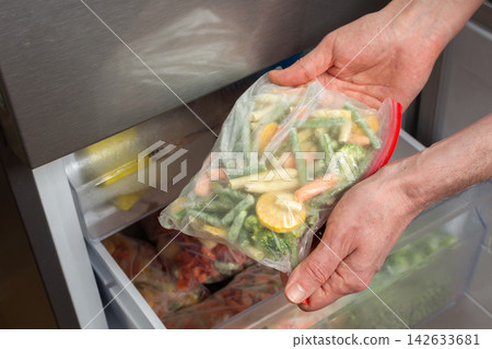 A man reaches into the refrigerator and takes out a bag of frozen vegetables. He is preparing to cook during the afternoon in a kitchen setting A man reaches into the refrigerator and takes out a bag of frozen vegetables. He is preparing to cook during the afternoon in a kitchen setting 142633681