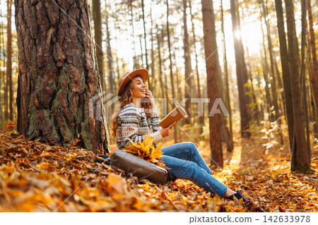 Pretty young woman in hat reading interesting book near tree in autumn forest. Enjoying nature Relax Pretty young woman in hat reading interesting book near tree in autumn forest. Enjoying nature Relax 142633978
