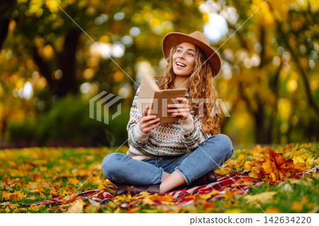Tourist is reading a book and enjoying the autumn landscape in the park while sitting on plaid. Tourist is reading a book and enjoying the autumn landscape in the park while sitting on plaid. 142634220