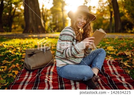 Tourist is reading a book and enjoying the autumn landscape in the park while sitting on plaid. Tourist is reading a book and enjoying the autumn landscape in the park while sitting on plaid. 142634225