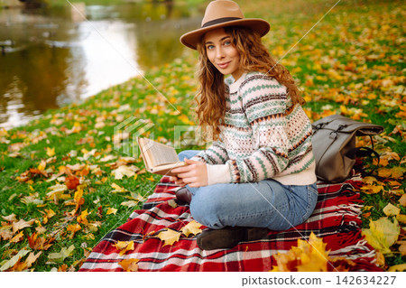 Tourist is reading a book and enjoying the autumn landscape in the park while sitting on plaid. Tourist is reading a book and enjoying the autumn landscape in the park while sitting on plaid. 142634227