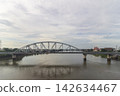 A steel arch bridge and elevated roadway cross an urban river beneath a broad cloudy sky with power lines and city buildings in the background. 142634467