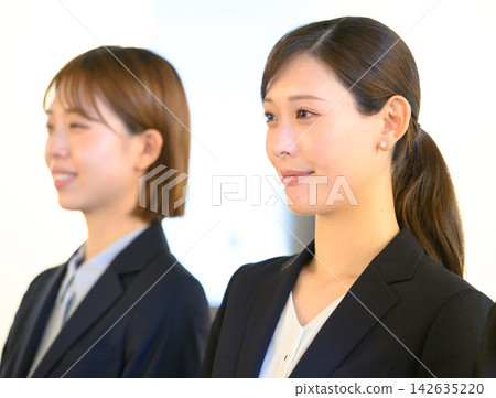 Two female employees standing side-by-side, looking straight ahead in a business setting. Two female employees standing side-by-side, looking straight ahead in a business setting. 142635220