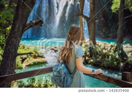 Woman traveler admiring El Chiflon waterfall from a wooden viewpoint in Chiapas Mexico with wooden railings tropical forest clear blue water nature travel and rear view. Woman traveler admiring El Chiflon waterfall from a wooden viewpoint in Chiapas Mexico with wooden railings tropical forest clear blue water nature travel and rear view. 142635408