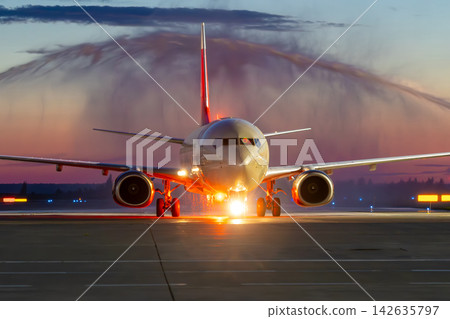 Front view of a white passenger airplane passing through a water arch at evening. First visit aircraft at the airport Front view of a white passenger airplane passing through a water arch at evening. First visit aircraft at the airport 142635797