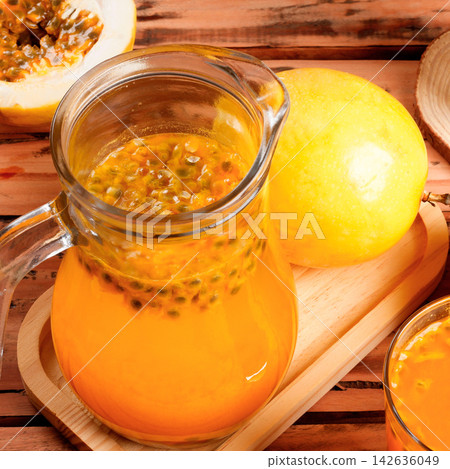 Refreshing tropical drink made with passion fruit, served in a pitcher and a glass, accompanied by whole and cut fruit on a rustic wooden table. 142636049